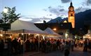 Ein belebter Marktplatz mit Verkaufsständen und vielen Menschen bei Nacht. Im Hintergrund steht ein Kirchturm, der hell beleuchtet ist.