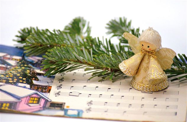 A decorative fabric Christmas angel stands on sheet music. Next to it are fir branches, and a festive winter landscape is visible in the background.