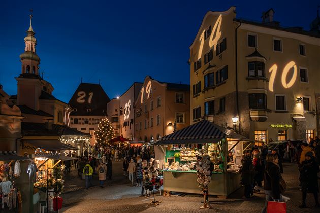 Ein lebhafter Weihnachtsmarkt mit festlich beleuchteten Ständen und zahlreichen Besuchern. Die Gebäude sind im Abendlicht erleuchtet und eine große Tanne steht im Zentrum.