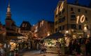 Ein lebhafter Weihnachtsmarkt mit festlich beleuchteten Ständen und zahlreichen Besuchern. Die Gebäude sind im Abendlicht erleuchtet und eine große Tanne steht im Zentrum.