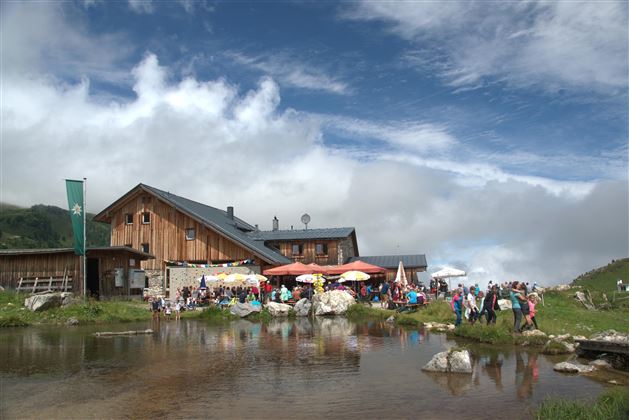 Une maison de montagne animée avec de nombreux visiteurs et des parapluies colorés. Au premier plan, il y a un petit étang et le ciel est nuageux.