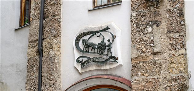 Ein dekoratives Schild aus Metall, das einen Wagen darstellt. Es ist an einer alten, steinernen Wand angebracht.