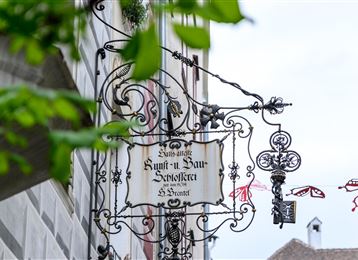 Un'insegna tradizionale di una locanda pende da un edificio. Sullo sfondo si vedono un cielo sereno e foglie verdi.