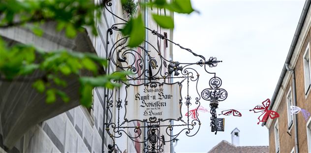 Ein traditionelles Schild eines Gasthauses hängt an einem Gebäude. Im Hintergrund ist ein klarer Himmel und grüne Blätter zu sehen.