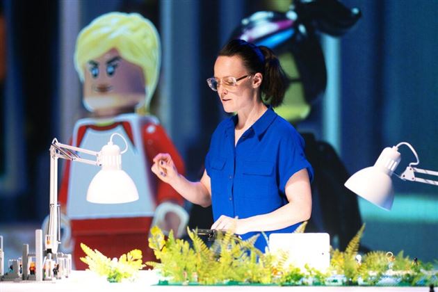 Une personne en chemise bleue se tient à une table avec des plantes et des lampes. En arrière-plan, de grandes figurines d'un système de construction sont visibles.