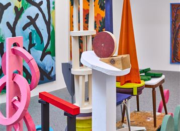 Une installation artistique avec des formes géométriques colorées et différents matériaux. En arrière-plan, on peut voir des fresques murales colorées.