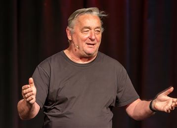 Ein Mann spricht auf einer Bühne und gestikuliert lebhaft. Er trägt ein schwarzes T-Shirt und wirkt freundlich und einladend.