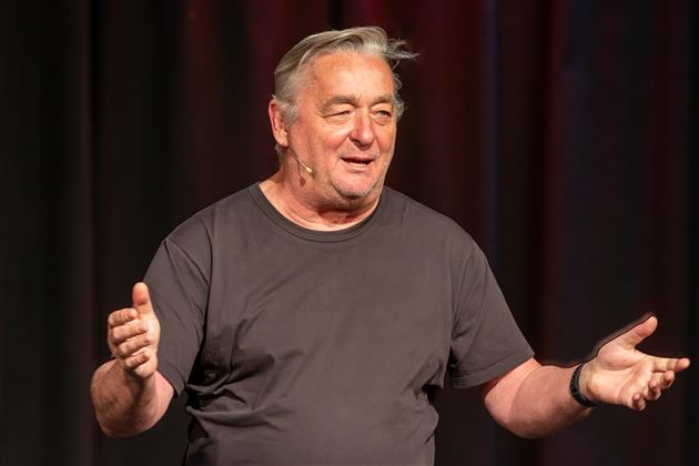 Un uomo parla su un palco e gesticola vivacemente. Indossa una maglietta nera e appare amichevole e accogliente.