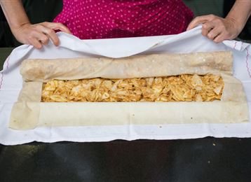 Una persona tiene un impasto con un ripieno di pezzi di mela. L'impasto viene arrotolato con cura e si trova su un panno bianco.