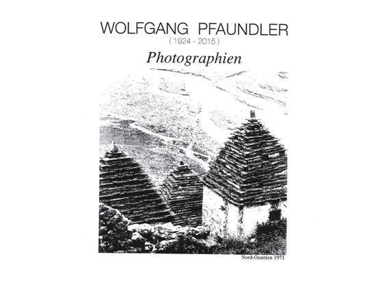 Eine Schwarzweißaufnahme von traditioneller Architektur mit geschlossenen Dächern aus Schindeln. Im Hintergrund sind Hügel und eine ländliche Landschaft sichtbar.