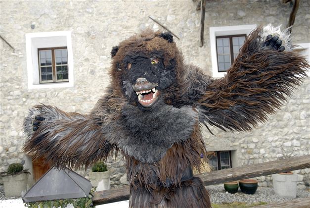 Ein bärähnliches Kostüm mit furchterregendem Gesicht und pelzigen Armen steht vor einem alten Gebäude. Der Hintergrund zeigt Fenster und eine steinerne Wand.