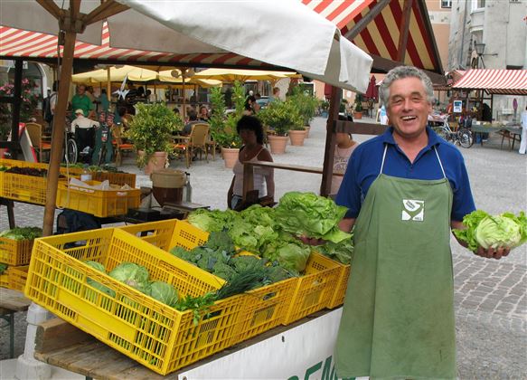 Een vriendelijke verkoper staat op een markt en houdt verse groenten in zijn handen. Op de achtergrond zijn meer groentekisten en marktkramen zichtbaar.