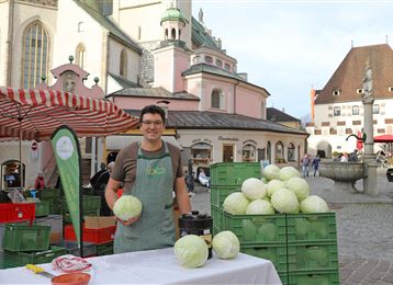 Ein Marktstand mit einem Verkäufer, der frische Weißkohlköpfe anbietet. Im Hintergrund sind historische Gebäude und ein Brunnen zu sehen.