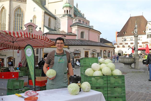 A market stall with a vendor offering fresh heads of white cabbage. In the background, historical buildings and a fountain can be seen.