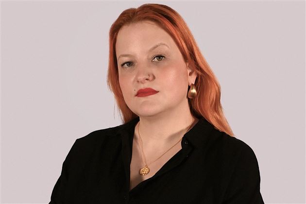 Une femme aux cheveux roux porte une chemise noire. Elle regarde directement l'objectif et a une expression confiante.