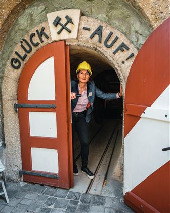 Une personne se tient à l'entrée d'une mine avec l'inscription « BONNE CHANCE ! ». Elle porte un casque jaune et regarde dehors.
