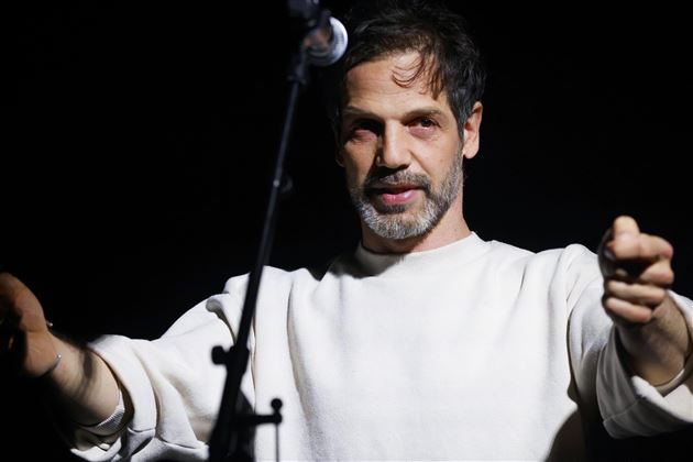 Un homme se tient sur une scène et parle dans un microphone. Il porte un pullover clair et a un regard intense.