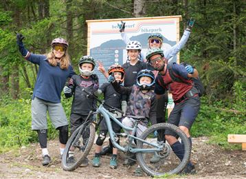 Un groupe de six personnes en tenue de cyclisme se tient dans une zone forestière. Ils posent à côté d'un VTT et s'amusent.