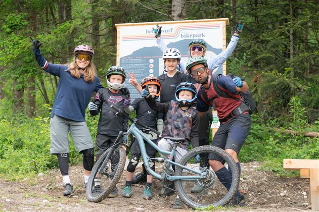 Een groep van zes personen in fietsoutfits staat in een bosgebied. Ze poseren naast een mountainbike en hebben plezier.
