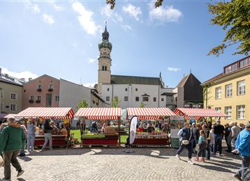 Ein lebendiger Marktplatz mit bunten Verkaufsständen und vielen Menschen. Im Hintergrund ist ein historischer Gebäudeturm zu sehen.