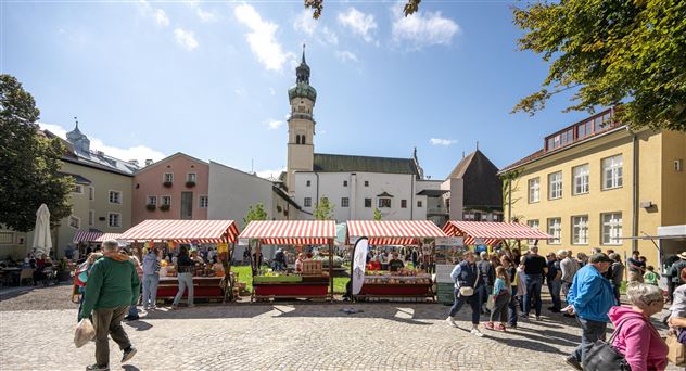 Ein lebendiger Marktplatz mit bunten Verkaufsständen und vielen Menschen. Im Hintergrund ist ein historischer Gebäudeturm zu sehen.