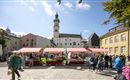 Ein lebendiger Marktplatz mit bunten Verkaufsständen und vielen Menschen. Im Hintergrund ist ein historischer Gebäudeturm zu sehen.
