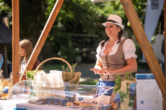 Eine Frau in traditioneller Tracht steht an einem Stand mit regionalen Produkten. Im Hintergrund ist ein grüner Garten zu sehen.
