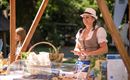 Eine Frau in traditioneller Tracht steht an einem Stand mit regionalen Produkten. Im Hintergrund ist ein grüner Garten zu sehen.