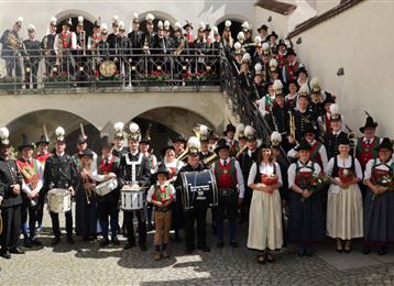 Eine große Gruppe von Menschen in traditioneller Kleidung steht vor einem Gebäude. Sie halten Instrumente und Blumen in den Händen.