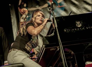 Una cantante si esibisce con passione al pianoforte sul palco. Sullo sfondo si possono vedere musicisti e uno sfondo colorato.