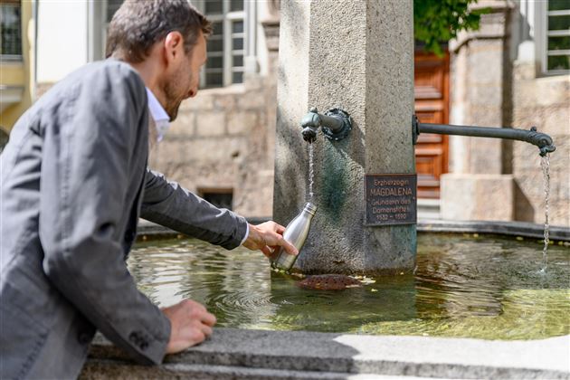 A man fills a bottle with water from a well. The well is made of stone and is located in an urban environment.