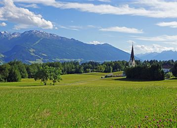 Un paysage pittoresque avec des prairies verdoyantes et une petite église. À l'arrière-plan, on peut voir des montagnes majestueuses et un ciel bleu.