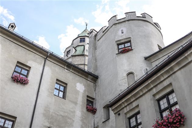 Eine alte Burg mit Zinnen und Fenstern, die mit Blumen geschmückt sind. Der Himmel ist teilweise bewölkt.
