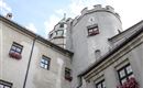 Eine alte Burg mit Zinnen und Fenstern, die mit Blumen geschmückt sind. Der Himmel ist teilweise bewölkt.