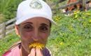 Eine Frau sitzt auf einer Wiese und hält eine Löwenzahnblüte vor ihr Gesicht. Im Hintergrund sind grüne Hügel und Kühe zu sehen.