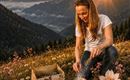 Eine Frau sitzt in einer blühenden Wiese und zündet etwas in einer Schale an. Im Hintergrund sind Berge und ein wunderschöner Sonnenuntergang zu sehen.