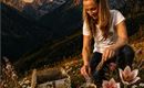 Eine Frau sitzt auf einer Blumenwiese in den Bergen und streut etwas aus einer Schale. Im Hintergrund leuchtet der Mond über den Gipfeln.