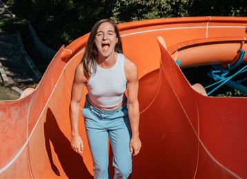 A woman is standing on a slippery water slide and laughing loudly. In the background, green trees can be seen.