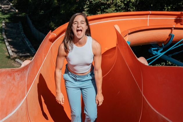 Une femme est debout sur un toboggan glissant et rit aux éclats. En arrière-plan, on peut voir des arbres verts.