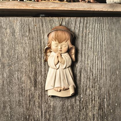 Un angelo con le mani giunte pende da una superficie di legno. La figura ha i capelli castani e indossa un habit bianco.
