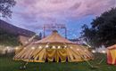 Een grote, gele circustent met kleurrijke gordijnen staat op een weide. De lucht is bedekt met paarse wolken, en lichten twinkelen aan de tent.