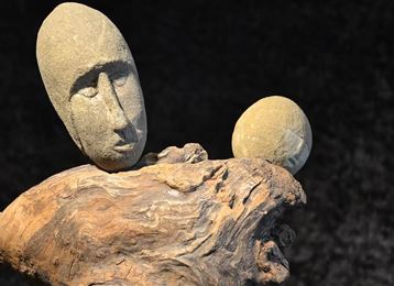 Une sculpture artistique en bois et en pierres, représentant des visages humains. Les figures semblent être assises sur un morceau de bois de racine.