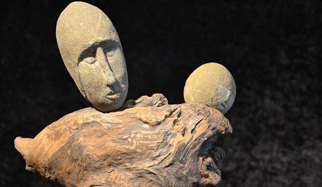 Una scultura artistica in legno e pietre che rappresenta volti umani. Le figure sembrano sedere su un pezzo di legno di radice.