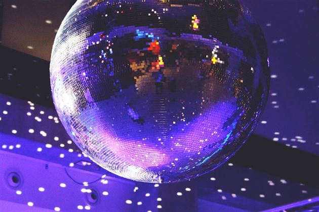 A disco ball hangs from the ceiling and reflects colorful beams of light. The background shows a relaxed party atmosphere with many colors.