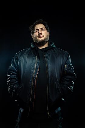 A man stands in a dark environment, wearing a black bomber jacket and has his hands in his pockets. His gaze is confident and directed straight at the camera.