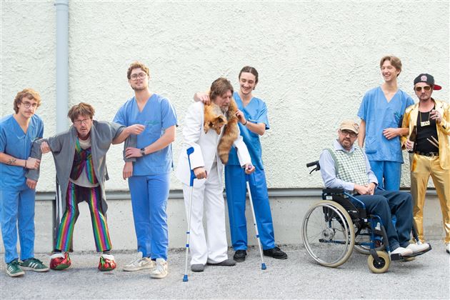 Eine Gruppe von jungen Männern in medizinischen Scrubs steht zusammen. Ein Mann im Rollstuhl und ein anderer mit einem Hund sind ebenfalls dabei.