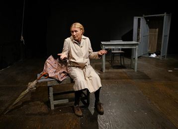 Une vieille femme est assise sur un banc et gesticule avec les mains. Dans la pièce, il y a une table et un coffre en bois, le décor est simple et sombre.