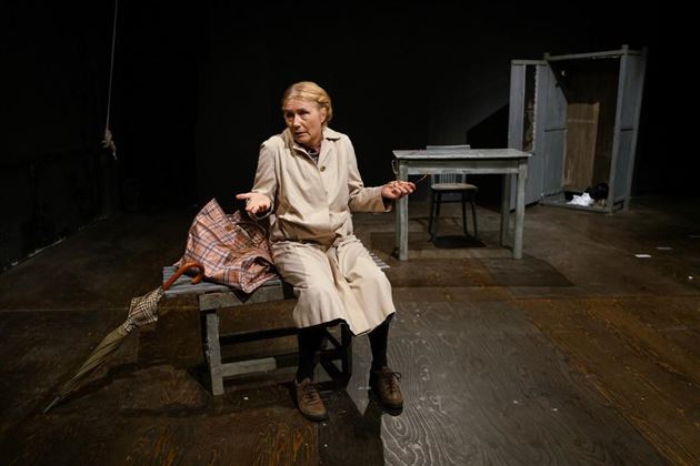 Een oudere vrouw zit op een bank en gebaart met haar handen. In de ruimte staan een tafel en een houten kist, de achtergrond is eenvoudig en somber.