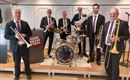 A six-member Dixieland band is standing on a stage. They are wearing suits and are happily posing with their instruments.