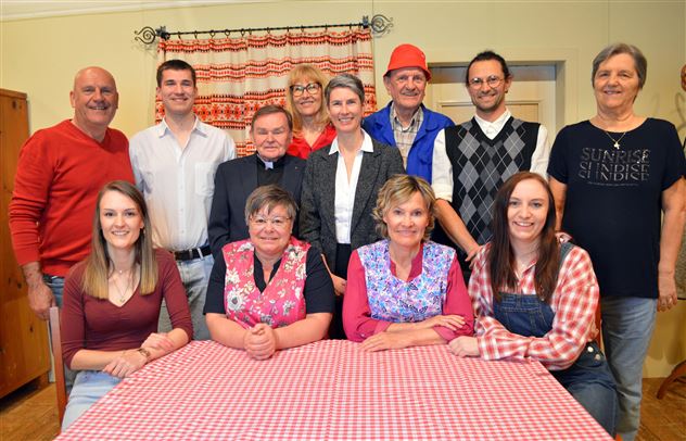 Un gruppo di persone è in piedi e seduto attorno a un tavolo con una tovaglia a scacchi. Ridono e sembrano trascorrere un momento felice insieme.